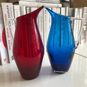 Puede incluir: Dos jarras de vidrio, una roja y otra azul, con un asa de vidrio transparente. Las jarras están sobre una superficie blanca.