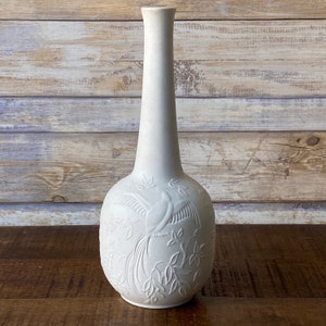 May include: White ceramic vase with an embossed design of a bird with a long tail and leaves. The vase has a tall, slender neck and a round body.