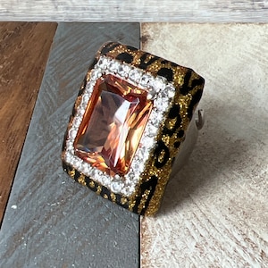 May include: A gold and black ring with a large, rectangular, orange-colored gemstone surrounded by clear rhinestones.