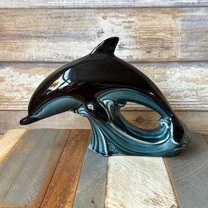 Può includere: Una statuetta di delfino in ceramica con corpo nero lucido e base a onda turchese. Il delfino è arcuato, come se saltasse dall'onda. La statuetta è esposta su una superficie di legno.