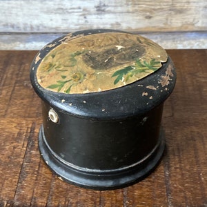 Könnte beinhalten: Eine kleine, runde, schwarze Holzkiste mit einem Blumendesign auf dem Deckel. Der Deckel ist leicht beschädigt und die Kiste hat ein kleines Loch an der Seite.