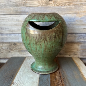 May include: A green ceramic vase with a unique cut-out design. The vase has a brown glaze with green streaks and a rounded base.