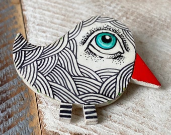 May include: A white ceramic bird with black and white striped patterns, a large blue eye, and a red beak. The bird has small black and white striped legs.