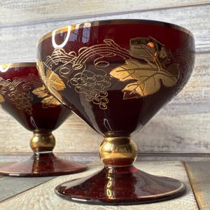May include: Two burgundy glass goblets with gold leaf accents. The goblets feature a grapevine design with gold leaves and grapes.