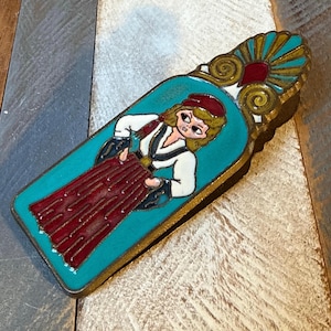 May include: A vintage decorative metal pin depicting a woman in traditional dress. The pin has a turquoise background with a gold border. The woman wears a red skirt, white blouse, and a red hat. The top of the pin features a gold and turquoise design.