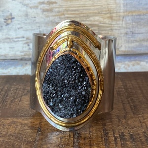 May include: A silver cuff bracelet with a gold-toned design featuring a black crystal in the center.