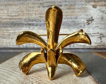May include: A gold-toned metal brooch in the shape of a stylized flower with five petals. The brooch has a textured surface and a pin on the back.