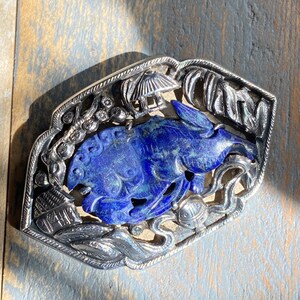 May include: A silver and blue lapis lazuli brooch featuring a stylised rabbit design. The rabbit is surrounded by intricate floral and geometric patterns.