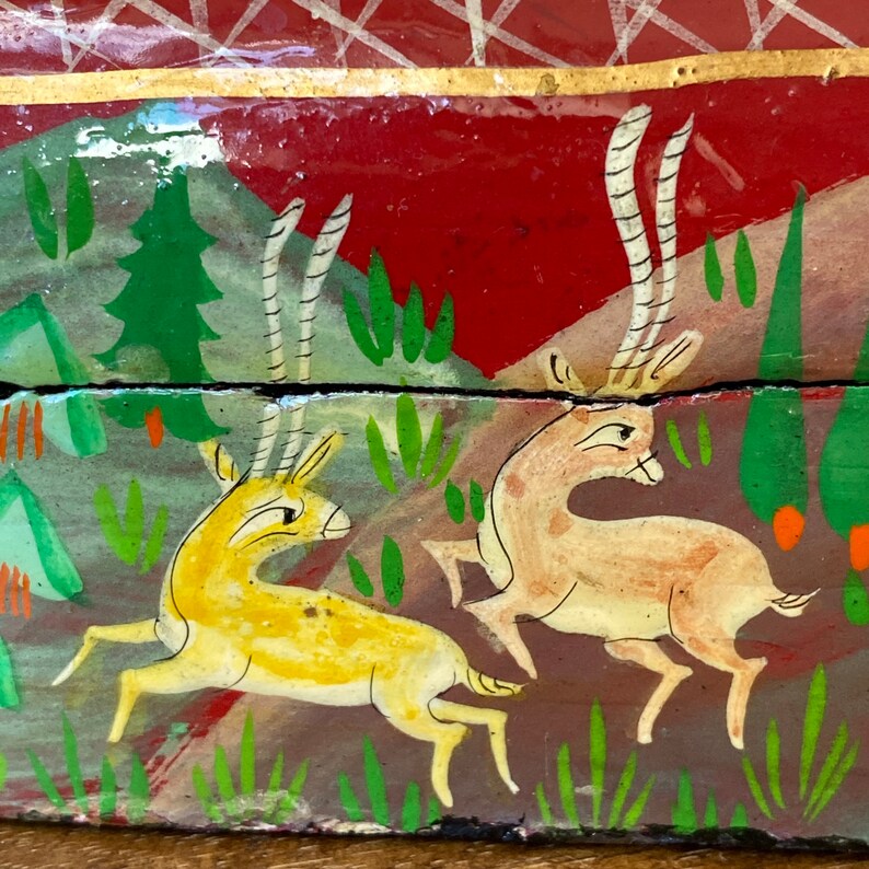Op de afbeelding: Handbeschilderde houten doos met een rode achtergrond en een sc&egrave;ne met twee herten in een bos. De herten zijn geschilderd in geel en bruin met witte geweien. Het bos is geschilderd in tinten van groen en bruin.