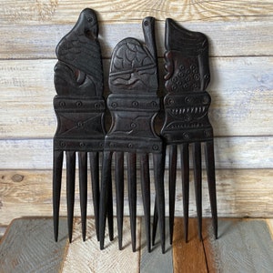 May include: Three wooden combs with intricate carvings. Each comb has a different design, including a bird, a geometric pattern, and a stylised human face. The combs are dark brown and have a polished finish.