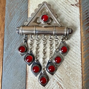 May include: A silver-toned pendant with a triangular top and a cylindrical bar. The pendant features seven red, circular gemstones set in diamond-shaped silver frames, suspended by chains. The design has an antique aesthetic.