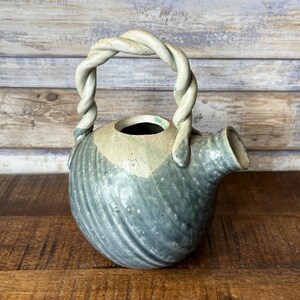May include: A ceramic teapot with a twisted handle. The teapot is primarily blue-green with a cream-coloured top and a spout. The handle is a light cream colour. The teapot has a round body and a textured surface.
