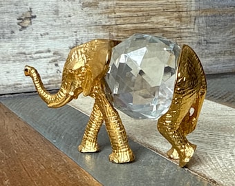 May include: A gold-colored elephant figurine with a textured surface, holding a large, clear, faceted crystal ball. The elephant has detailed features and stands on four legs. The background is a mix of wood and gray-toned surfaces.