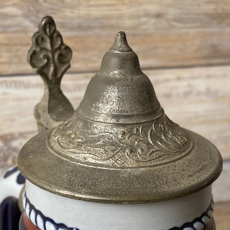 Pu&ograve; includere: Primo piano di un coperchio in peltro per una boccale di birra. Il coperchio &egrave; decorato con un intricato motivo floreale e ha una piccola maniglia decorata.