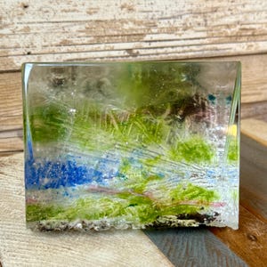 Puede incluir: Un bloque de arte de vidrio rectangular con un diseño de paisaje. La obra de arte presenta una mezcla de colores verdes, azules y blancos, que se asemejan a una escena natural. El vidrio es transparente, lo que permite ver los colores a través del bloque.