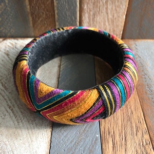 Könnte beinhalten: Ein farbenfrohes Stoffarmband mit einem geometrischen Muster in Rot-, Orange-, Gelb-, Lila-, Blau-, Schwarz- und Goldtönen. Das Armband besteht aus einem weichen, gewebten Stoff und hat ein schwarzes Samtfutter.