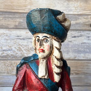 Puede incluir: Una figurita de madera pintada de una persona que lleva un sombrero azul, una chaqueta roja y azul y una peluca blanca. La figurita está pintada con un estilo realista y tiene una cara detallada.