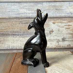 May include: A glossy black wolf figurine in a howling pose. The sculpture has detailed carving, with visible teeth and a raised head. The wolf is seated, with its front paws extended. The background is a mix of wooden planks.