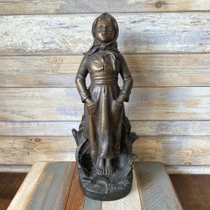 May include: A bronze sculpture of a young girl wearing a long dress and a headscarf. The sculpture is standing on a base with a floral design and the inscription "Enfant de la Ville".
