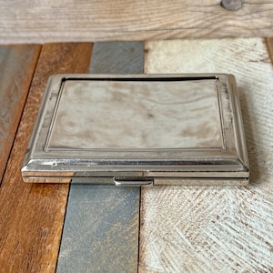 May include: A rectangular, silver-toned metal case with a hinged lid. The case has a smooth, reflective surface and a simple, elegant design. The background is a wooden surface with various colors.