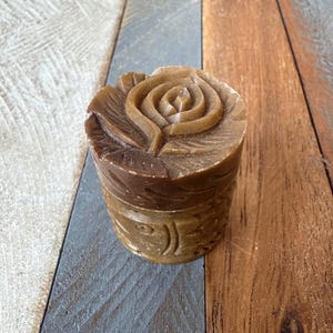 May include: A brown, cylindrical container with a carved rose design on the lid. The sides of the container feature a carved floral pattern. The item is made of a solid, possibly stone-like material.