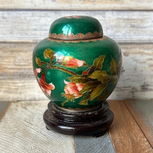 May include: A green cloisonné jar with a floral design and a wooden base. The jar has a lid with a gold trim.