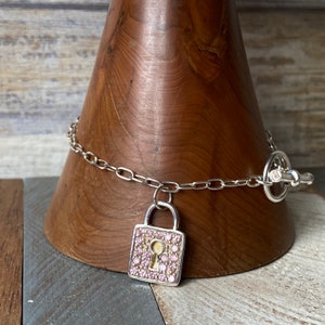 Puede incluir: Un collar de cadena de plata con un dije de candado de pedrería rosa. La cadena está unida con un cierre de plata.