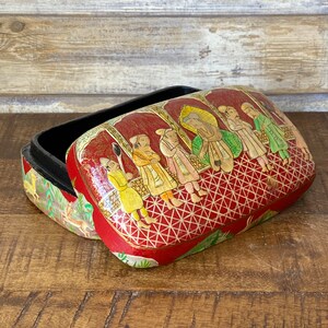 Op de afbeelding: Een rode papier-mach&eacute; doos met een zwart interieur en een kleurrijk handgeschilderd tafereel van mensen in traditionele kleding. De doos is versierd met een geometrisch patroon.