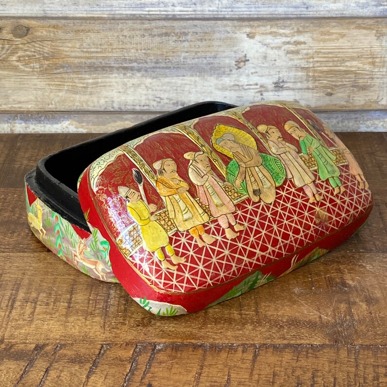 Op de afbeelding: Een rode papier-mach&eacute; doos met een zwart interieur en een kleurrijk handgeschilderd tafereel van mensen in traditionele kleding. De doos is versierd met een geometrisch patroon.