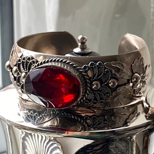 Könnte beinhalten: Silberfarbenes Armreif-Armband mit einem ovalen, facettierten, roten Edelstein. Das Armband hat aufwendige Blumen- und Perlendesigns. Das offene Design ermöglicht einfaches Tragen. Ein dekoratives Schmuckstück.