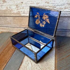 May include: A rectangular glass box with a hinged lid, constructed with dark metal solder joints. The box features deep blue glass panels, and the interior base is mirrored. The lid displays a gold-colored botanical leaf and flower decal.
