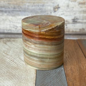 May include: A cylindrical stone box with a lid. The box is made of layered onyx, displaying bands of green, brown, and tan hues. The box is sitting on a wooden surface.