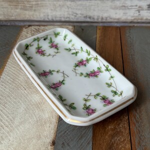 Puede incluir: Un pequeño plato de porcelana blanca con borde dorado y un diseño floral de rosas rosas y hojas verdes.
