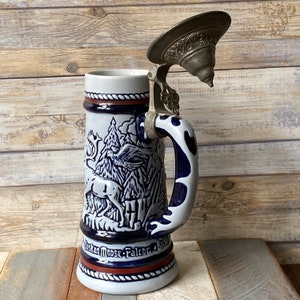 Pu&ograve; includere: Una boccale di birra in ceramica bianca con un design blu e rosso che presenta un alce, un falco e altri animali selvatici. La boccale ha un coperchio in metallo con un design decorativo.