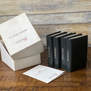 Puede incluir: Una caja blanca con la palabra "CONCORDE" y "British Airways" impresas, junto a cuatro libros negros con las inscripciones "Italiano", "Alemán", "Español" y "Francés". También hay un folleto con la misma marca.