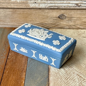 May include: A rectangular blue trinket box with white relief detailing. The box features floral and classical figures in relief. The lid is adorned with flowers and a central relief scene. The box is set against a wooden background.