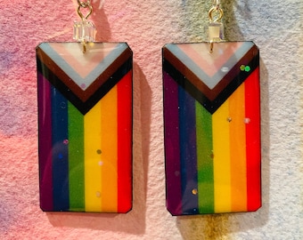 Könnte beinhalten: Ein Paar rechteckige Ohrringe mit dem Progress Pride Flaggen-Design. Die Ohrringe haben einen Regenbogenverlauf mit schwarzen, braunen, hellblauen, rosa und weißen Streifen. Silberfarbene Fischhaken-Ohrdrähte und kleine transparente Perlen vervollständigen das Design.