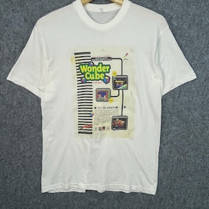May include: A white short-sleeve t-shirt featuring a graphic print on the chest. The print displays the text Wonder Cube, Japanese characters, and three small rectangular images showing game scenes, alongside a black and white striped vertical bar.