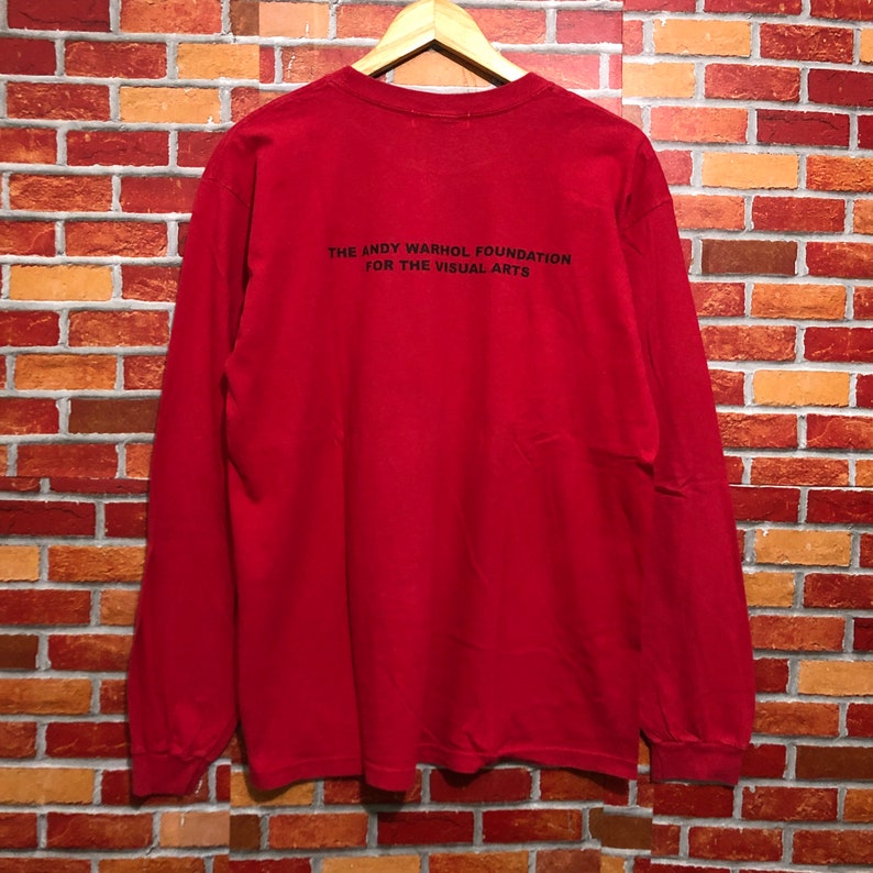 Puede incluir: Una camiseta roja de manga larga con el texto "THE ANDY WARHOL FOUNDATION FOR THE VISUAL ARTS" impreso en negro en la espalda. La camiseta est&aacute; colgada en una percha de madera contra una pared de ladrillos.