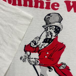 May include: White t-shirt featuring a red and black illustration of a gentleman with a cane and red coat. The text "Johnnie Walker" is printed in red at the top of the shirt.