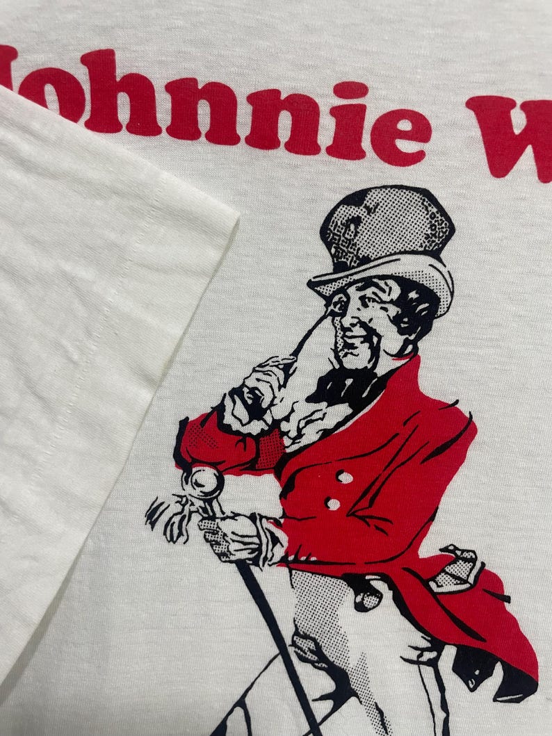May include: White t-shirt featuring a red and black illustration of a gentleman with a cane and red coat. The text "Johnnie Walker" is printed in red at the top of the shirt.