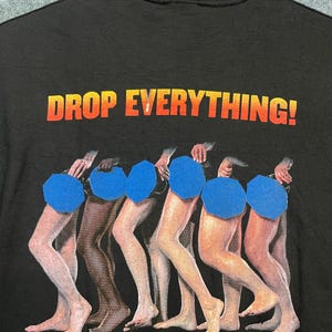 以下が含まれることがあります： 黒いTシャツで、黄色とオレンジ色の文字で「DROP EVERYTHING!」と書かれています。その下には数組の裸足の脚のグラフィックがあり、それぞれの脚の付け根部分は青い八角形の図形で部分的に隠されています。