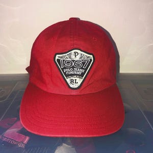 May include: A bright red baseball cap featuring a central shield-shaped patch. The patch is black with white text that reads 1967, Polo Jeans Company, Ralph Lauren, and RL, accompanied by a small letter P and wing-like graphic elements.