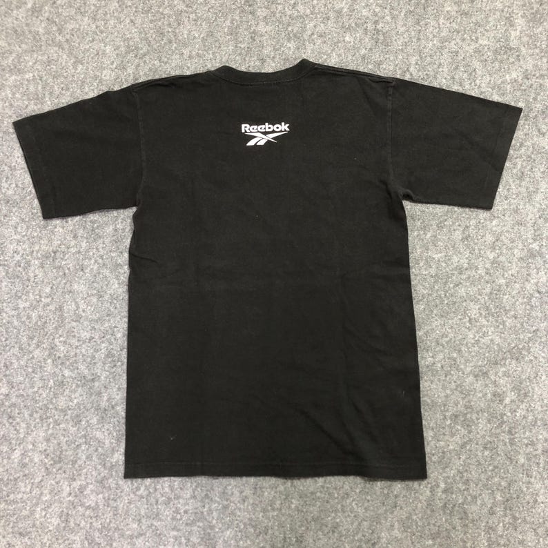 Puede incluir: Camiseta negra con el logo blanco de Reebok en la parte superior de la espalda. La camiseta tiene mangas cortas y cuello redondo. La tela parece ser de algod&oacute;n o una mezcla de algod&oacute;n.