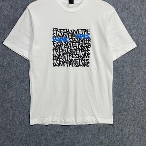 Pode incluir: Uma t-shirt branca de manga curta com um gráfico quadrado no peito. O gráfico consiste em linhas repetidas da palavra FRESH em texto preto, com uma instância da palavra FRESH destacada em azul, tudo numa fonte de estilo graffiti.