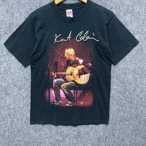 Puede incluir: Una camiseta negra de manga corta con un estampado gráfico de Kurt Cobain tocando la guitarra acústica en un escenario. El texto "Kurt Cobain" está impreso en blanco sobre la imagen. La camiseta tiene cuello redondo y una etiqueta de marca Anvil.