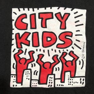Puede incluir: Camiseta negra con un dise&ntilde;o gr&aacute;fico cuadrado blanco. El dise&ntilde;o presenta las palabras "CITY KIDS" en letras rojas, con tres figuras rojas con los brazos levantados sobre un paisaje urbano. El dise&ntilde;o es al estilo de Keith Haring.