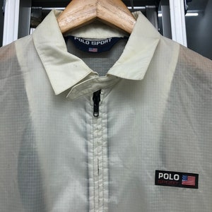 May include: Off-white Polo Sport jacket with a full-length zipper and a collared neckline. The jacket features the Polo Sport logo and an American flag patch on the chest. The jacket is hanging on a wooden hanger.