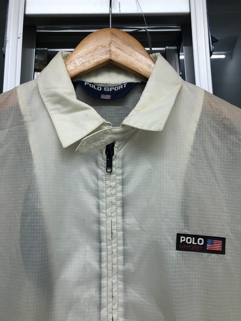 May include: Off-white Polo Sport jacket with a full-length zipper and a collared neckline. The jacket features the Polo Sport logo and an American flag patch on the chest. The jacket is hanging on a wooden hanger.