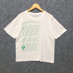 May include: White t-shirt with green text and a graphic. The text is a statement about corporate responsibility. The t-shirt is displayed on a wooden hanger.